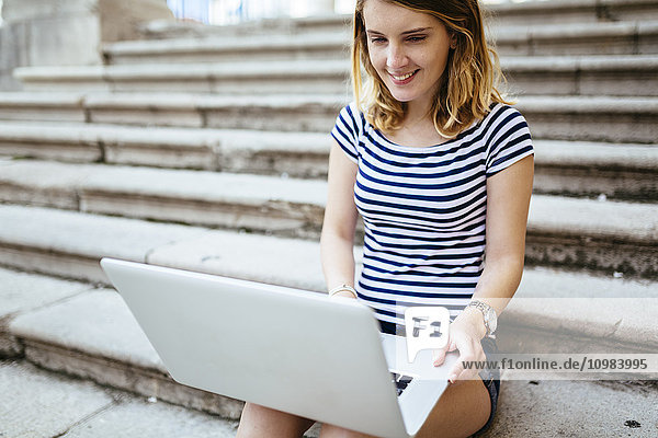 Lächelnde junge Frau sitzt auf der Treppe mit Laptop