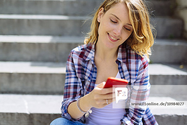 Porträt einer lächelnden jungen Frau  die mit dem Smartphone auf der Treppe sitzt.