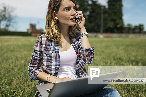 Young woman with laptop sitting on a meadow telephoning with smartphone