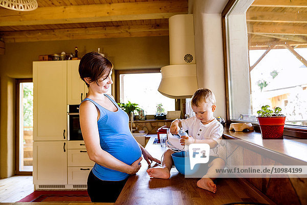 Kleiner Junge hilft seiner schwangeren Mutter in der Küche mit elektrischem Schnurrbart