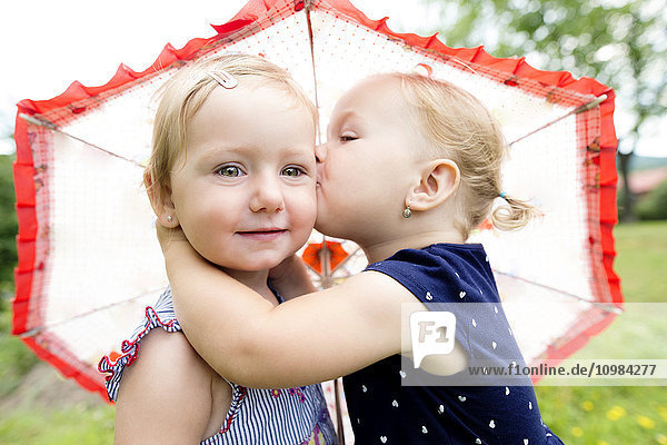 Kleines Mädchen  das seine Schwester unter einem Regenschirm küsst.
