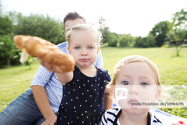 Kleines Mädchen mit Vater und Schwester in einem Park  das dem Betrachter ein Croissant anbietet.