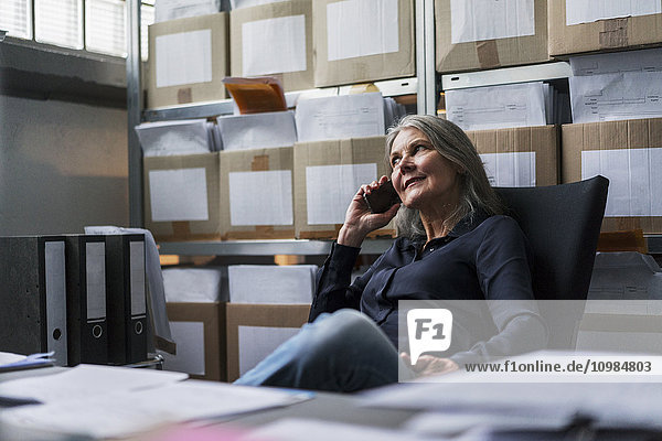 Seniorin sitzt auf einem Bürostuhl in einer Fabrik und telefoniert mit dem Handy.