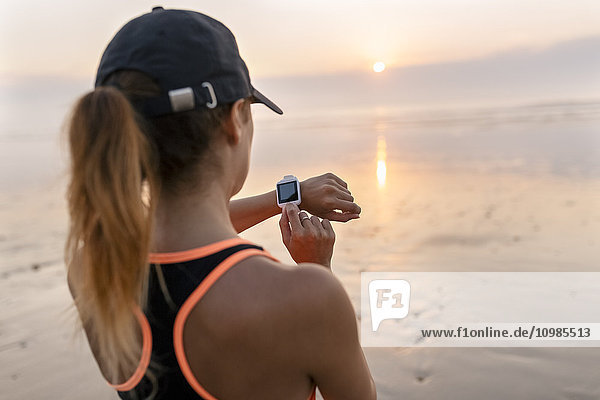 Junge Sportlerin mit Blick auf die Smartwatch am Strand bei Sonnenuntergang