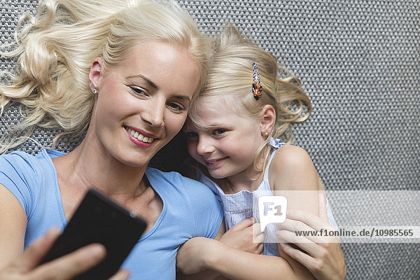 Mutter und ihre kleine Tochter liegen auf dem Boden und nehmen Selfie mit dem Smartphone.