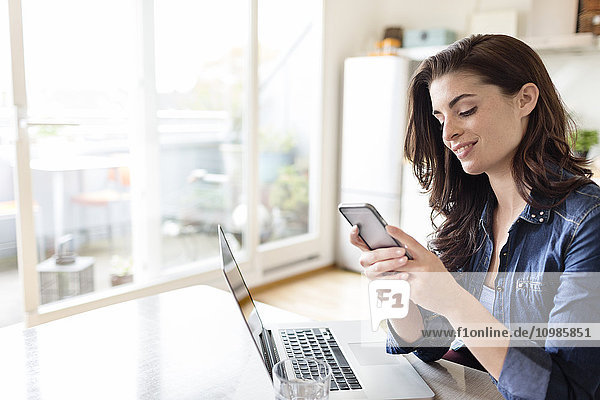 Smiling young woman at home using cell phone and laptop