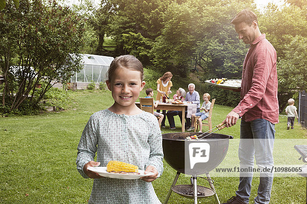 Lächelndes Mädchen hält einen Teller mit Maiskolben auf einem Familiengrill.