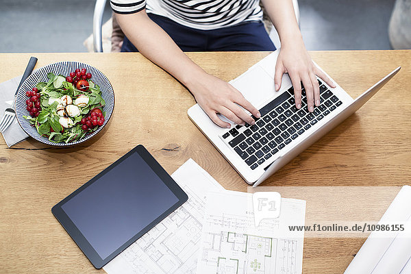 Frau am Schreibtisch mit Laptop neben Bauplan und Salat