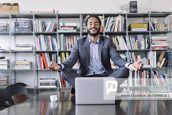 Ein junger Geschäftsmann sitzt im Büro und meditiert vor dem Laptop.