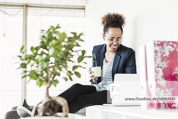 Lächelnde Geschäftsfrau mit Tasse Kaffee auf dem Laptop