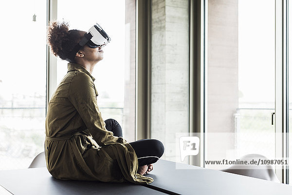 Frau im Büro sitzt auf einem Konferenztisch und trägt eine Virtual-Reality-Brille.
