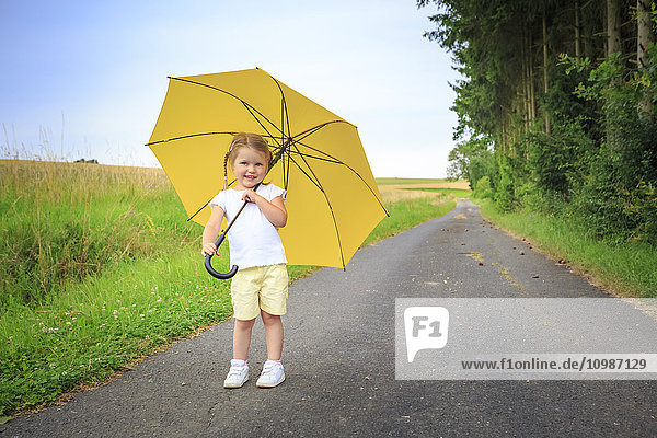 Smiling little girl with yellow umbrella standing on country road