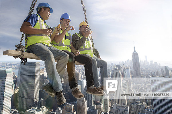 Three construction workers sitting on suspended scaffolding high above city having lunch