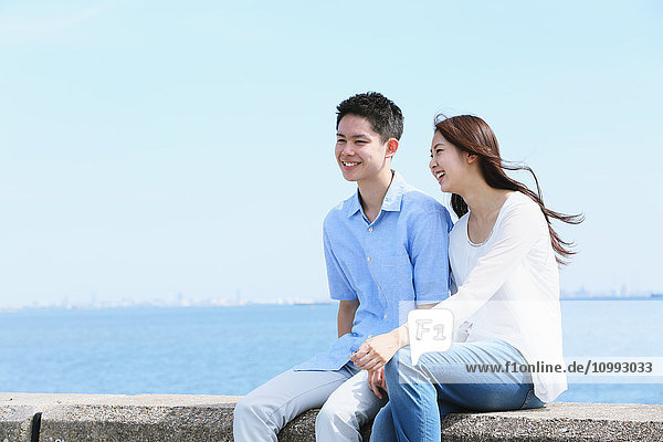 Junges japanisches Paar sitzt auf einer Betonmauer am Meer