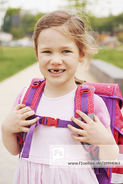 Porträt eines 5-jährigen Schulmädchens mit rosa und lila Schultasche  das lächelnd in die Kamera schaut und zeigt  dass ihr die Vorderzähne fehlen