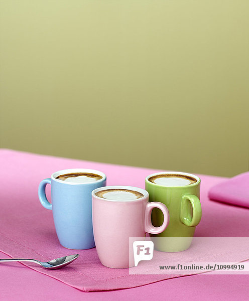 Trio von pastellfarbenen Demitasse-Tassen mit Espresso und Milchschaum auf rosa Tischset mit Löffel
