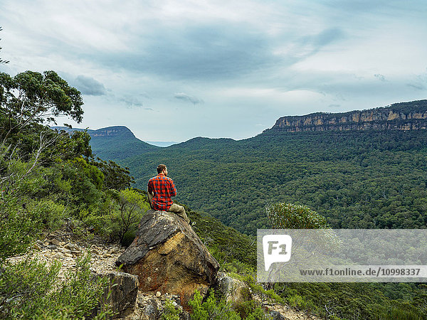 Australien  New South Wales  Katoomba  Rückansicht eines mittelgroßen Mannes  der auf einem Felsen sitzt und auf die Blue Mountains blickt