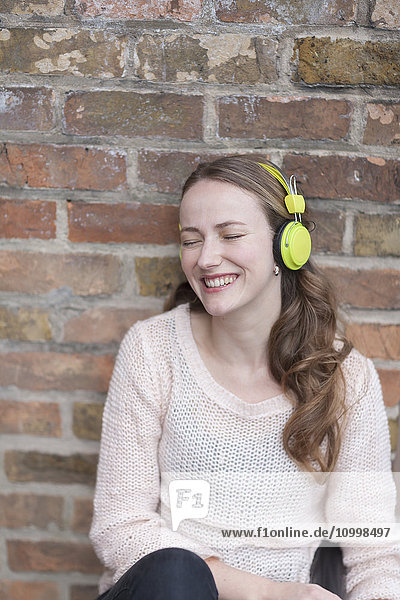 Mid-adult woman sitting and leaning against brick wall with closed eyes and headphones on