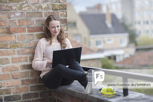 Mid-adult woman sitting on balcony railing and using laptop