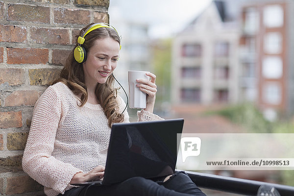Mid-adult woman with headphones on sitting on balcony railing holding coffee cup and using laptop