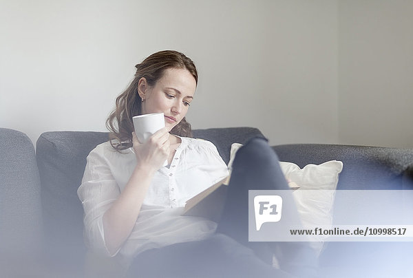 Frau im mittleren Erwachsenenalter sitzt mit Buch und Tasse Kaffee auf dem Sofa