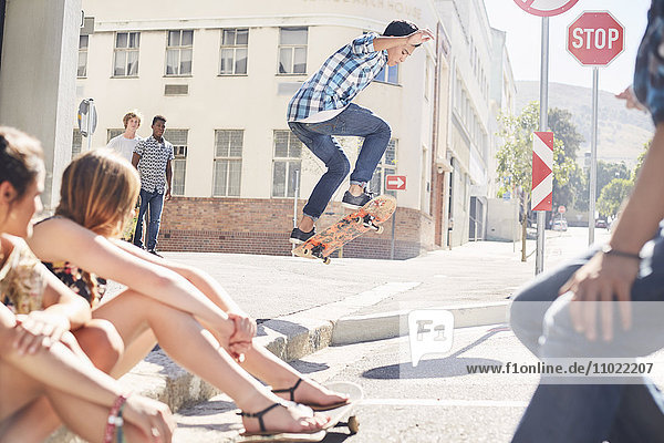 Freunde beobachten Teenager-Junge beim Skateboard-Springen an der sonnigen urbanen Ecke.