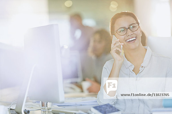 Lächelnde Geschäftsfrau im Gespräch mit Handy am Computer im Büro