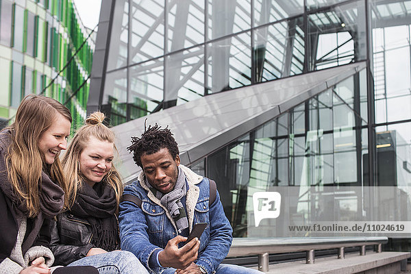 Friends looking at smart phone while sitting against glass building