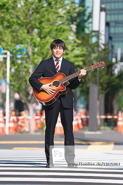 Junger japanischer Geschäftsmann mit Gitarre am Zebrastreifen