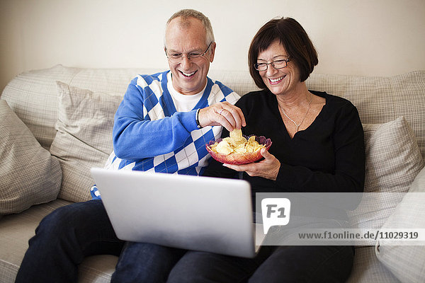 Älteres Paar auf dem Sofa sitzend  Laptop schauend  Schale mit Chips haltend