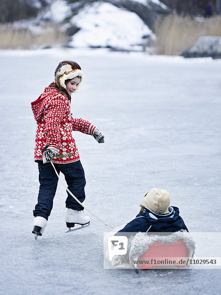 Mädchen  das seinen Bruder beim Schlittschuhlaufen auf einem zugefrorenen See auf einem Schlitten zieht
