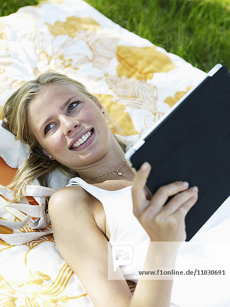 Junge Frau liegt auf einer Decke und hält ein digitales Tablet  lächelnd