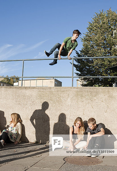 Jugendlicher springt über Geländer  während andere Jugendliche an der Wand sitzen