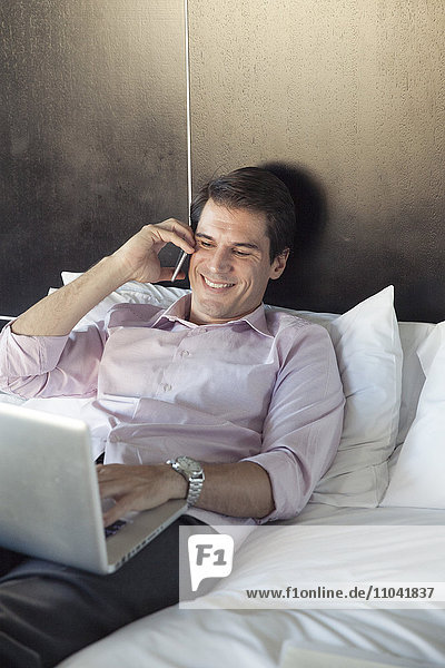 Mann mit Laptop und Handy im Bett