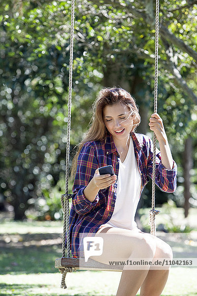 Young woman sitting on park swing  text messaging with smartphone