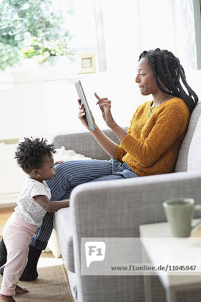 Schwarze Frau benutzt digitales Tablet auf dem Sofa neben ihrer kleinen Tochter