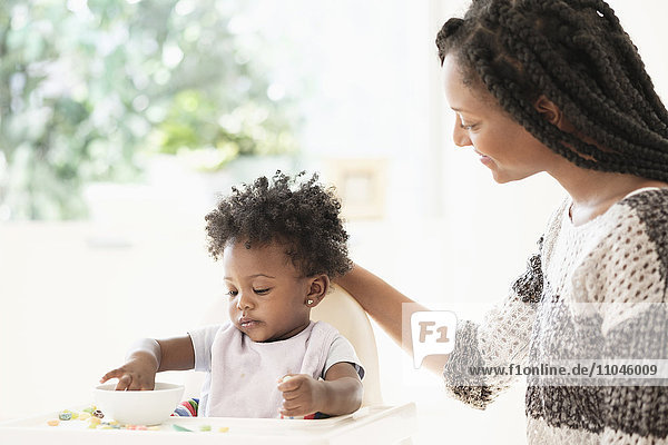 Schwarze Frau beobachtet ihre kleine Tochter beim Müsliessen im Hochstuhl