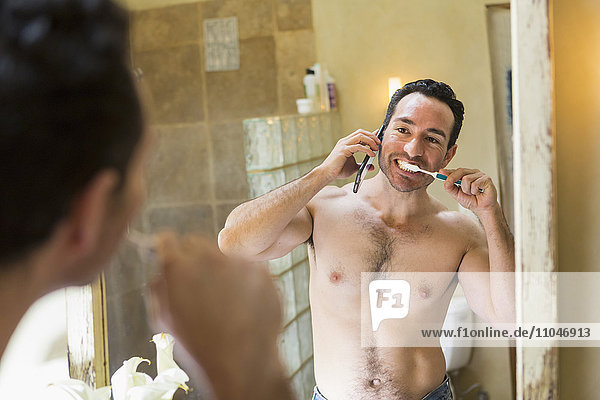 Hispanic man using cell phone and brushing teeth in mirror