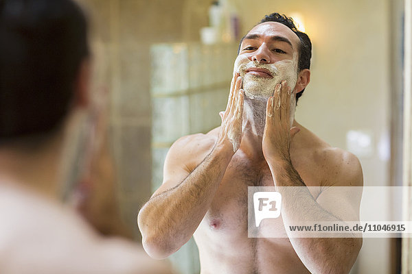 Hispanic man applying shaving cream to face in mirror