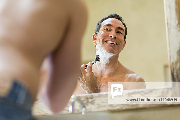 Hispanic man using razor to shave face in mirror