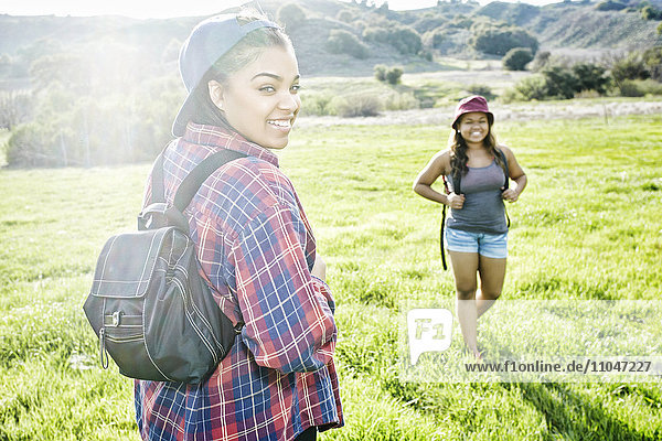 Smiling Mixed Race sisters backpacking in field near mountain