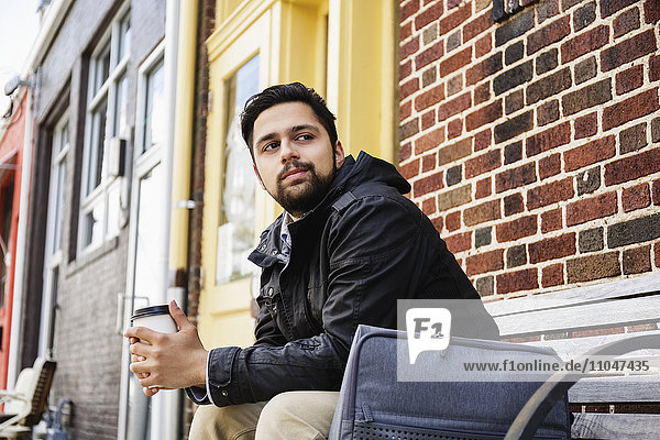 Hispanic man sitting on city bench drinking coffee