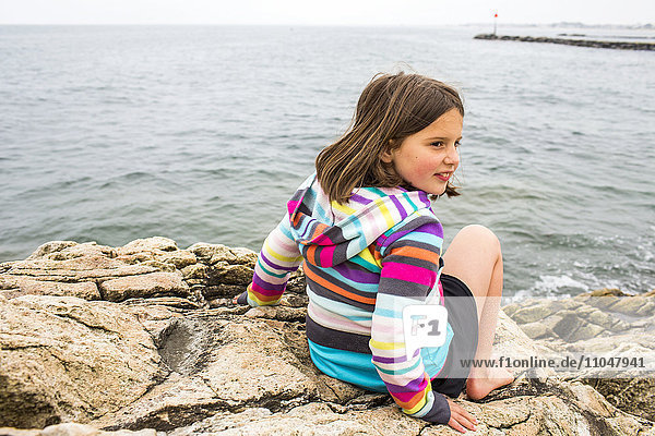 Smiling Caucasian girl sitting on rocks at ocean