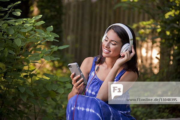 Lächelnde hispanische Frau  die im Hinterhof mit Kopfhörern ihr Handy benutzt