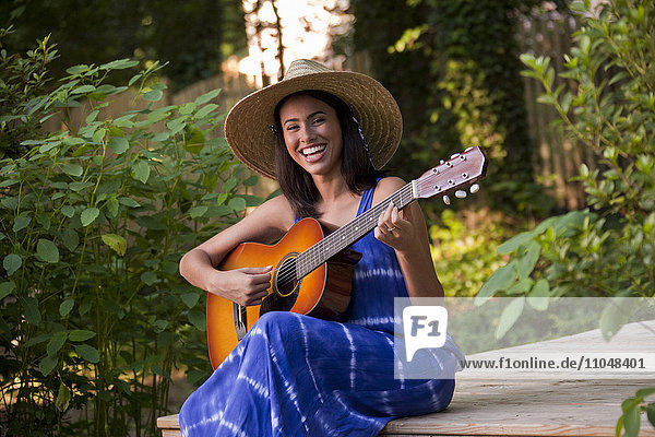 Lachende hispanische Frau spielt Gitarre auf der Terrasse im Hinterhof