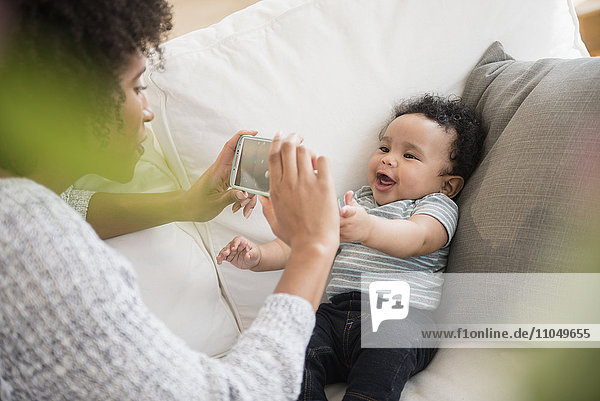 Mutter fotografiert Babysohn mit Handy auf Sofa