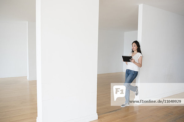 Hispanic woman leaning on wall of empty apartment holding digital tablet