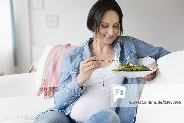 Schwangere kaukasische Frau isst Salat auf dem Sofa