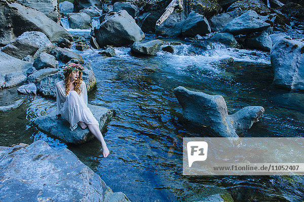 Kaukasische Frau mit Blumenkrone auf einem Felsen am Fluss