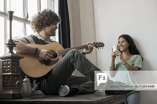 Loft living. A young man playing guitar to an appreciative audience  a young woman holding a smart phone and taking a photograph.